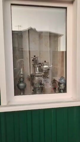 This media file displays a close-up view of a window with a reflection. Through the glass, various objects are visible, including a large, ornate silver samovar, a silver teapot, a kerosene lamp, and two smaller decorative vases. The items appear to be displayed on a shelf inside a room, with a curtain visible behind them. The reflection in the window shows an outdoor scene, with a building and some sky.

The exterior of the building is also visible, featuring green corrugated metal siding below the window. To the right of the window, a section of a log cabin is visible, with decorative flowers in a tall vase placed in front of it. The overall setting suggests an outdoor space adjacent to a dwelling. The lighting in the reflection and the general ambient light suggest it might be daytime, possibly overcast. There is no visible text in the media file. The arrangement of the objects inside the window hints at a display or collection.