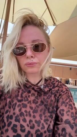 A woman with blonde hair, sunglasses, and a leopard-print hoodie is taking a selfie outdoors, possibly by a swimming pool. The sun is shining, creating a bright atmosphere. Behind her, lounge chairs and tables are visible, suggesting a resort or hotel setting. The woman's expression is neutral, and she appears relaxed. The image doesn't contain any visible text. The context of Abu Dhabi, United Arab Emirates, places the scene in a warm, likely tropical environment, consistent with the poolside setting and bright sunlight. The woman's attire, a hoodie, might suggest it's not extremely hot at the moment, or she's seeking some shade and comfort from a light breeze. The overall impression is one of leisure and relaxation in an outdoor vacation setting.