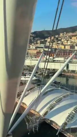 From an elevated perspective, possibly from within a large structure, the video shows a panoramic view of a harbor. The harbor is filled with various boats and yachts docked at piers. In the distance, the coastline is lined with buildings and hills, with a clear blue sky overhead dotted with a few clouds. The framing of the shot includes structural elements in the foreground, painted in white and designed with a radial arrangement, which partially obstruct the view. The overall impression is one of a calm and sunny day at a bustling port city.