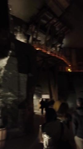 The video shows a group of people walking through a dark, cavernous space. The space is lit by artificial lights that illuminate the walls and a wooden walkway. The walkway is elevated and has a railing. A sign on the wall reads "KOMORA ERASMA BARACZA.  WIZEB WIEKU."  The people are walking in the same direction. The camera follows behind them, focusing on the back of their heads and shoulders. The environment looks like a mine or a cave.