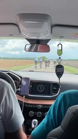 The video, shot from inside a car, shows a group of cyclists riding on a paved road. The car appears to be following the cyclists, maintaining a distance. Inside the car, two people are visible, one in the driver's seat and another in the passenger seat. The dashboard features a car stereo with a navigation system and a phone mounted on a holder. A radio device is hanging from the rearview mirror. The scenery outside the car consists of fields and trees under a cloudy sky. The cyclists are wearing colorful racing attire. The car's interior is partially visible, including the gear shift and controls.