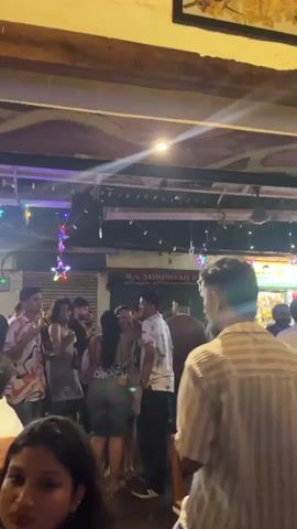 The video shows a crowded outdoor seating area of a restaurant or bar with people standing, talking, and dancing. There are string lights with star decorations hung overhead. Some people are holding drinks. The camera pans right, showing people seated at tables. The background features a painted mural on the wall. The overall atmosphere is lively and festive.