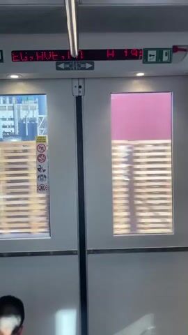 The video shows the interior of a train with two sets of doors. A digital display above the doors shows the time as 19:34 and the temperature as 23 degrees Celsius. The windows of the doors show a blur of passing scenery, indicating the train is in motion. On the left door window, there are several warning signs. The right door window shows more of the passing scenery. A portion of a person's head is visible in the bottom left corner of the frame.