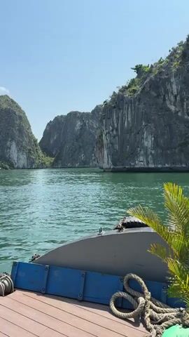 The video shows a view from a boat cruising through Ha Long Bay, Vietnam. The camera pans to the left, showcasing the green water and the limestone karsts that jut out of the bay. The boat has a wooden deck with a winch and rope visible. The sky is blue with a few scattered clouds.