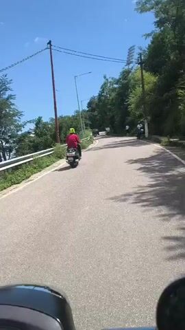 A first-person perspective shot shows a person riding a scooter on a paved road. The road curves to the right and is bordered by a grassy embankment on the left and a tree-lined hill on the right. Several other scooters are visible on the road ahead, some with riders, and one parked on the side. The sky is blue with scattered clouds, and the sun is shining. The scooter's handlebars and mirror are visible in the foreground.