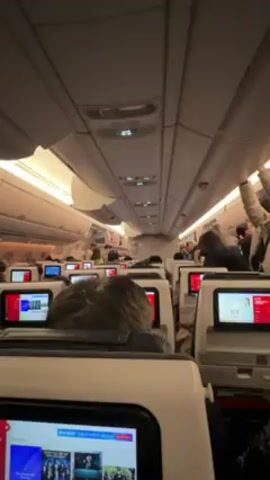 The interior of an airplane cabin is shown from the perspective of someone seated in a middle row. Overhead, the cabin ceiling is a light off-white with closed luggage compartments and several illuminated signs. There are two rows of seats visible on either side of the aisle, with a small screen attached to the back of each seat. The screens display various images. People are seated throughout the plane, and some are standing up to access the overhead compartments. The overall lighting in the cabin is soft and warm.