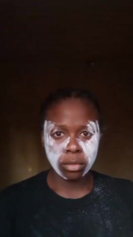 A person with dark skin is applying a white, creamy substance to their face and neck. The substance is primarily concentrated around their eyes, nose, mouth, and neck, leaving patches of skin visible. The person is wearing a black shirt and stands against a dark background. They rub the substance into their skin, then open their mouth wide in a surprised expression.