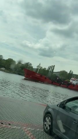 The video shows a gray car parked on a ferry with a red tanker ship passing by. The tanker, named "ALEXANDRION" in white letters, is moving to the left of the frame on a body of water. The sky is overcast with a mix of light and dark clouds. Trees line the shore in the background. The ferry's surface is textured with a diamond plate pattern and painted with red and gray stripes. The car's front is visible, including a portion of the hood and a front tire.