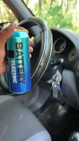 A person's hand holds a blue and teal can of "BATTERY ELECTRIC" energy drink inside a car. The car's interior is visible, with the dashboard and part of the door. The open car door reveals a sun-dappled forest scene with trees and green foliage outside. The camera then pans to show the exterior forest environment more fully.