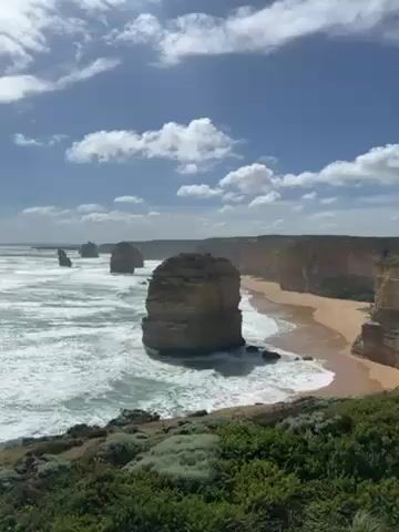 The video shows a scenic coastal view under a bright blue sky with scattered white clouds. In the foreground, green shrubbery covers the edge of a cliff. Beyond the cliff, the ocean waves crash against several large, rugged rock formations standing in the water. A sandy beach stretches along the base of steep, layered cliffs on the right side of the frame. The strong wind creates whitecaps on the ocean surface. The sound of wind is prominent in the video.