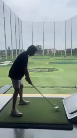 The video depicts a person, likely a young male, playing golf at what appears to be a Topgolf or similar venue. He is dressed in a black shirt, light grey shorts, and white sneakers. He is standing on a green mat and swings a golf club, hitting a golf ball into the distance. The background shows multiple circular targets on a green field, surrounded by safety netting and tall metal poles. The sky is overcast. A digital interface or screen is visible to the right of the person.