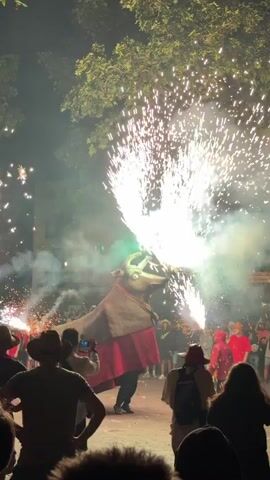 The video shows a nighttime outdoor event with a large bull effigy that appears to be part of a performance or parade. The effigy is adorned with a brown cape and a red skirt. It is positioned in the center of the frame, with a crowd of people gathered around it. The effigy is actively discharging fireworks from its head and hindquarters, creating large bursts of white and green sparks and smoke. The crowd consists of individuals of various ages, many holding up phones to record the spectacle. The environment is dimly lit, with trees visible in the background and some architectural elements suggesting a street or public square. The atmosphere appears festive and exciting, with the fireworks being the main focal point of the activity.