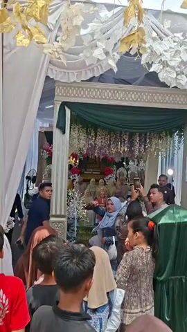 The video shows a formal gathering, likely a wedding ceremony, taking place indoors under a decorated tent. In the background, a group of people, possibly family members or guests, are seated on a raised platform adorned with flowers, white artificial trees with lights, and elaborate drapes. The individuals on the platform are dressed in traditional attire, some wearing headscarves. The bride, wearing a distinctive headdress and traditional clothing, is prominently seated. In the foreground, a crowd of people, including children, are gathered, some holding up mobile phones to capture the event. A woman wearing a blue headscarf appears to be gesturing and speaking to someone in the crowd. The overall atmosphere suggests a celebratory occasion with an emphasis on tradition and documentation.