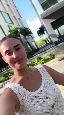 A light-skinned woman with her hair pulled back, wearing a white crocheted tank top and a delicate necklace, is standing outside and filming herself. The sun is bright. She is looking directly at the camera and slightly smiling. Behind her, there are palm trees and buildings with a mix of white, yellow, and orange colors. The camera moves slightly as she holds it.