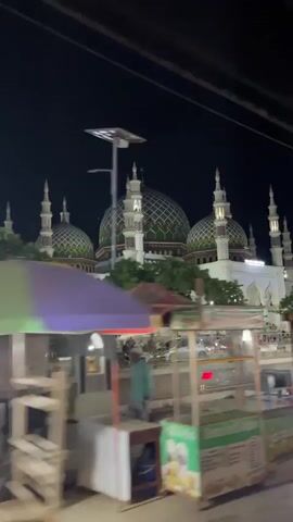 The video shows a mosque at night, with its domes and minarets illuminated. In the foreground, there are several food carts and people walking by. The overall scene suggests a busy street outside a mosque during evening hours.