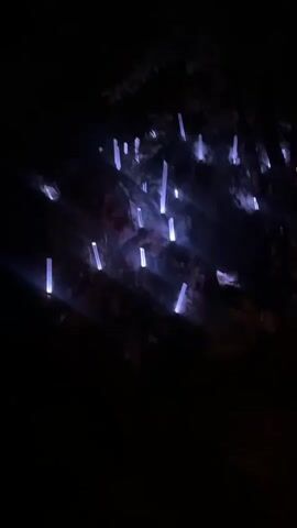 The video shows an outdoor nighttime scene with illuminated objects and a person. Initially, the camera pans across a dark, rocky surface with many vertical, rectangular lights emitting a purple glow. These lights are scattered and appear to be embedded in or attached to the rock face. The camera then moves to show a wooded area at night. More of the purple rectangular lights are visible, some are in clusters and others are more spread out amongst the trees. The lighting in the woods is varied, with green and red lights also illuminating the foliage and ground. In the foreground, a person, appearing to be male, is standing behind a DJ setup. He is wearing a white t-shirt and is interacting with the equipment. The DJ setup is bordered by a bright orange, wavy neon light. The overall impression is of an outdoor art installation or an electronic music event held in a natural setting.
