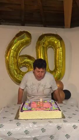 A woman is celebrating her 60th birthday in a room with white walls and a wooden ceiling. She wears a gray t-shirt and stands behind a table covered with a plastic tablecloth. On the table is a large rectangular birthday cake decorated with pink and purple frosting and a "60" candle. Behind her are two large gold foil balloons forming the number "60" and several regular balloons in bronze, white, and black colors. The woman blows out the candles on the cake and smiles as people applaud. She then puts her hand to her eye as if wiping away a tear.