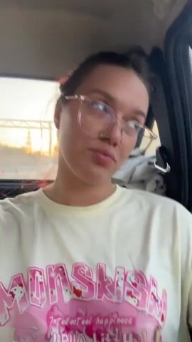 A young woman with her hair pulled up and wearing glasses and a white t-shirt is in the passenger seat of a vehicle. The t-shirt has a large pink graphic with the word "MONSSISIH" in large letters. Below this, in smaller letters, it reads "Intellectual happiness." The woman is making a pouting face. The scene appears to be taking place during the day, possibly late afternoon or early evening, given the lighting outside the vehicle, which shows a hazy, possibly sunset sky with a power line pylon in the distance. The setting appears to be a road in a rural or semi-rural area, suggested by the power line and the open landscape visible through the window. The woman's expression suggests a playful or lighthearted mood.