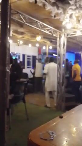 The video depicts a dimly lit indoor space, possibly a bar or restaurant, with multiple people present. The flooring appears to be artificial grass in sections. The ceiling is adorned with a grid of metal beams and what appears to be decorative cloud-like material. Some people are milling around, and one person in a light gray outfit raises his hands in the air, appearing to be celebrating or reacting to something, as cheering can be heard. The atmosphere suggests a festive or celebratory occasion. A pool table is in the foreground. The background includes a dimly lit area with blue lighting, possibly a separate part of the establishment.
