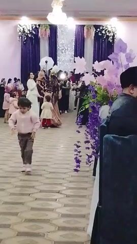 The video captures a wedding reception or party in a large hall. The floor is decorated with a geometric tile pattern, and the background is adorned with purple drapes, white floral arrangements, and a sparkling silver centerpiece. A chandelier hangs from the ceiling, providing bright overhead light. A small child in a pink sweater and grey pants walks across the floor, while other children are also scattered around. A woman in a long, peach-colored gown with an elaborate headpiece appears to be a central figure, possibly the bride, and is surrounded by other guests taking pictures with their phones and cameras. To the right of the frame, a table is decorated with large purple and pink flower arrangements, and a man sits at the table, partially obscured. Overall, the scene conveys a celebratory atmosphere with people mingling and taking photos at the festive event.