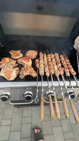 The video shows a close-up shot of a barbecue grill with various meats cooking. On the left side of the grill are several pork chops, some with grill marks and a light brown sear. To the right of the pork chops are multiple skewers filled with cubed meat, likely lamb or beef, which are also cooking and browning. Smoke is rising from the grill, indicating the cooking process is active. The grill itself appears to be a stainless steel gas grill with visible control knobs on the front. The background shows a paved patio area, and a person's feet wearing black sandals are visible at the bottom of the frame, suggesting someone is standing in front of the grill. The overall scene depicts an outdoor grilling event.