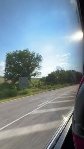 The video shows a view from the window of a moving vehicle, likely a bus, looking out at a rural landscape. The time of day appears to be late morning or early afternoon, given the bright sunshine and blue sky with scattered white clouds. The weather is clear and sunny.

The scene depicts a paved road with white lane markings, stretching into the distance. On either side of the road, there are green fields and trees. The trees vary in type, with some appearing as leafy deciduous trees and others as more conical evergreens. In the background, there are fields that appear to be cultivated or have been harvested.

The sun is shining brightly, casting a lens flare on the window. The reflection of the interior of the vehicle is also visible on the window, with a hint of a dark gray seatback and a pinkish headrest on the right side of the frame.

The overall impression is one of travel through a pleasant, sun-drenched countryside. There are no people visible in the frame. The activity taking place is the motion of the vehicle through the landscape.

There is some faint, vertically oriented text visible on the upper right portion of the window, near the edge. It is difficult to read clearly due to the glare and resolution, but it appears to be a serial number or identification marking, possibly related to the vehicle or the window itself. It looks like "20122" or a similar sequence.

The location context provided is Balgarene, Bulgaria. This suggests the scene is taking place in Bulgaria.