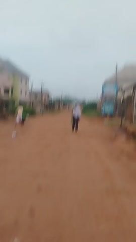 The video shows a dirt road in a village. There are several buildings on either side of the road, some of which appear to be residential. There are also some trees and vegetation visible. The sky is overcast. There are a few people walking on the road. One person is riding a bicycle. The video is shaky and the focus is not always sharp, suggesting it was filmed handheld.