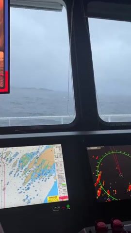 The video shows a view from inside a boat's cabin. Two large screens are visible in the foreground. The screen on the left displays a nautical chart with detailed information about landmasses and water depths. The screen on the right shows a radar display, indicating various targets with red and green markers, and a red line pointing ahead. The screens are mounted on a dark dashboard. Behind the dashboard, through a large window, the choppy, grey sea is visible under an overcast sky. Wipers are attached to the window. In the upper portion of the view, a multi-panel display shows four different video feeds. One feed shows a woman being interviewed in what appears to be an outdoor setting with a crowd in the background. Another feed shows people celebrating with red banners. A third feed shows two people's torsos, one wearing a red and white striped shirt, and the other a green shirt. The fourth feed displays a stadium with a scoreboard showing "0:38:05" and text in a foreign language. The overall lighting suggests a dim, possibly stormy day.