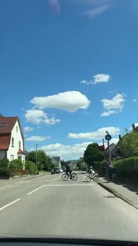 The video shows a street scene on a sunny day with a bright blue sky and scattered white clouds. In the foreground, the road is visible with white pedestrian crossing lines. Three people on bicycles are crossing the street. The person on the left is an adult wearing a dark shirt and pants, riding a dark bicycle. In the center, a child wearing a helmet and a light-colored shirt and shorts rides a small white bicycle. To the right, an adult wearing a red and black checkered shirt and dark pants rides a dark bicycle. There is a traffic light visible on the right side of the road, which is currently red, and a sign with a bicycle symbol. Houses and trees line the street on both sides. A yellow vehicle, possibly a bus, is visible in the distance. The perspective suggests the video is being recorded from inside a vehicle.