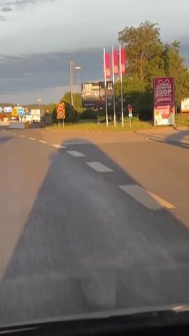 The video captures a forward-facing view from a vehicle approaching and passing through a border crossing at what appears to be the Slovenia-Italy border near Monrupino / Repentabor. The time of day is late afternoon or early evening, indicated by the warm, golden light and long shadows cast by the vehicle on the asphalt road. The sky is partly cloudy.

Initially, the scene shows a road with white dashed lines and a checkerboard pattern. A prominent yellow rectangular sign with a red border displays "POLICIJA POLICE" at the top and a circular speed limit sign with "10" inside, below which are silhouettes of a car and a truck. This suggests a 10 km/h speed limit for all vehicles, including trucks, at a police checkpoint. In the background, a large billboard reads "PARK POSTOJNSKA," likely advertising Postojna Cave Park in Slovenia. Red flags on poles are visible near the billboard.

As the vehicle moves forward, white concrete road barriers appear on the left side, delineating lanes or a controlled area. A blue square sign with white arrows is also visible in the distance, indicating directions.

Further ahead, a large blue square sign with "ITALIA" written in white surrounded by yellow stars (representing the European Union) marks the entry into Italy. Below this sign, smaller signs read "X814," "ANAS RA14," and "inizio competenza" (start of jurisdiction). On the right side of the road, a yellow concrete bollard and a red circular sign with a white horizontal bar and a diagonal red line (indicating a prohibition, likely 'No Entry' or similar restriction) are visible. The scene depicts a smooth transition across what was once a more formal border, now likely a routine entry point within the Schengen Area, still marked with national identifiers and some controls.