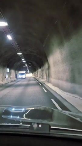 The video shows a first-person view from inside a vehicle driving through a tunnel. The tunnel walls are rough and appear to be made of rock or concrete. Lights are mounted along the ceiling of the tunnel, illuminating the road ahead. The road is paved and has white lane markings. In the distance, the end of the tunnel is visible, with natural light shining through. To the left, a white camper van is also driving through the tunnel. The dashboard of the vehicle is visible in the foreground.