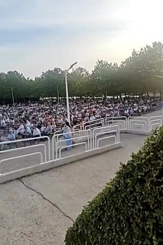 A wide shot captures an outdoor amphitheater filled with a large audience seated in rows of chairs. The audience members are diverse in age and attire, with many facing forward towards an unseen stage. In the foreground, a white metal railing forms a barrier. Behind the railing, a woman in a blue patterned dress stands and appears to be looking towards the seated crowd. Trees line the background, and a cloudy sky is visible above. The overall impression is of a public gathering or performance taking place in a park-like setting.