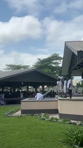 The video shows an outdoor gathering under a cloudy sky. In the foreground, a grassy lawn leads to a raised platform with a pool. On the platform, three individuals in white clothing stand near the pool, while another person in white is partially submerged in the water. In the background, two covered structures are visible. The left structure is larger and appears to house a crowd of people seated. The right structure, smaller, is open-sided and seems to serve as a covered area. Trees and buildings are visible beyond the structures. The overall scene suggests a religious or ceremonial event taking place outdoors.