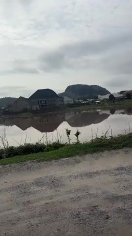 The video displays a flooded residential area in Angwan Sayawa, Nigeria, under an overcast sky. The murky brown floodwaters are extensive, reflecting houses and a distant, large hill or mountain.

The initial view captures several houses, some appearing large and modern with dark roofs, partially submerged or surrounded by water, their detailed reflections visible on the water's surface. A dirt road and some green vegetation occupy the immediate foreground.

The camera then pans across the scene, revealing more of the widespread flooding impacting numerous residential structures. The final shot focuses on a specific large, white house featuring a dark roof and multiple windows. The floodwater reaches close to its foundation, clearly reflecting the building. A concrete barrier or wall stands partially submerged in front of the house, and some debris floats on the water's surface. An air conditioning unit is visible on the side of the house.

No people are present in the video, and there is no discernible text. The scene depicts the impact of significant flooding on the community's infrastructure and landscape, under a cloudy daytime sky.