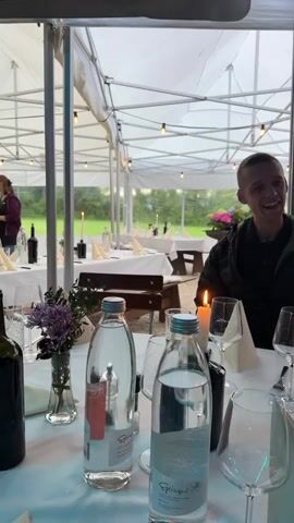 The video shows an outdoor gathering at tables set with white tablecloths and place settings. In the foreground, two bottles of water, wine glasses, and a lit candle are visible on a table. A young man in a dark jacket smiles at the camera. In the background, other people are seated at tables and interacting. The setting appears to be a restaurant or event space with a white tent-like covering overhead. The overall atmosphere is casual and social.