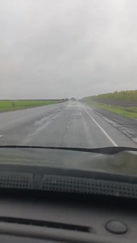 The video depicts a view from inside a car driving on a wet highway during a rainy day. The windshield wipers are in operation, clearing the rain. The road stretches into the distance, with fields on either side. Another car is visible ahead, driving in the same direction. The sky is overcast and gray.