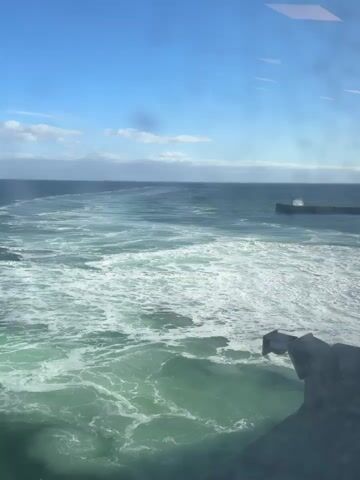 A video shows a body of water with waves and whitecaps under a blue sky with scattered clouds. A dark, angular object is visible in the foreground, partially obscuring the view of the water. In the distance, a structure that appears to be a breakwater or pier with a small building on it is visible in the ocean. The water is a greenish-blue color, and the waves are choppy. The sky is clear and bright.