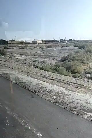 The video captures a view from a vehicle traveling on a paved road. The landscape is arid, with sparse vegetation and dusty terrain. In the distance, several industrial buildings or structures are visible. At approximately 00:01, a blue road sign with white lettering indicating "Zirabud" and an arrow pointing left appears. Later, at around 00:04, a triangular warning sign with a red border and a white interior is seen. The weather appears clear with a bright sky.