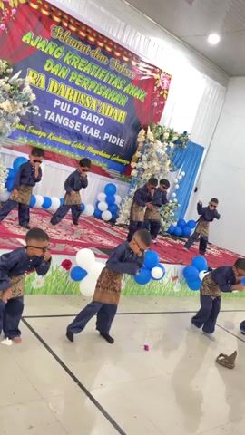 This video shows a group of young boys performing a dance on a stage decorated with balloons and a banner. The boys are all dressed in matching navy blue outfits with gold sashes and sunglasses. They appear to be performing a choreographed routine, with some boys in the front row bending down while others in the back row stand and gesture. The stage is set up in what appears to be an indoor venue, possibly a school or community hall, with white curtains in the background. A large banner at the back of the stage reads "SELAMAT DAN SUKSES AJANG KREATIFITAS ANAK DAN PERPISAHAN RA DARUSSA' ADAH PULO BARO KEC. TANGSE KAB. PIDIE". Below this, smaller text reads "Semoga Kesuksesan Selalu Menyertai Selamanya". The overall atmosphere seems festive and celebratory, likely marking an end-of-year event or graduation for the children from RA Darussa' Adah school.
