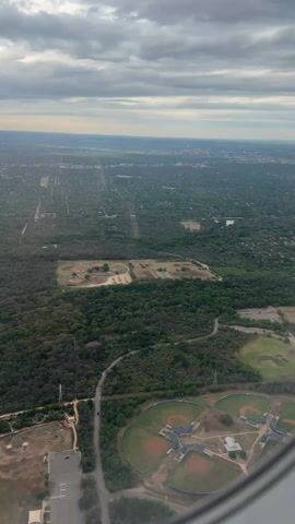 The video shows an aerial view of a landscape from an airplane window, including baseball fields, park areas, trees, roads, parking lot, and a distant skyline. The weather is cloudy. The view appears to be over a suburban or urban area with many trees and green spaces. The airplane engine comes into view.