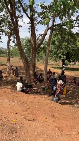 The video shows a group of people gathered under a tree in a rural area. There are adults and children, some sitting, some standing. The ground is reddish dirt. There are some buckets and other containers scattered around. The scene suggests a community gathering, perhaps for rest and water in a warm climate. The setting appears to be in a less developed country. The background shows trees, a clear sky, and a field.