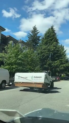 The video shows a small, four-wheeled forklift driving down a residential street in a suburban area on a sunny day with blue skies and scattered clouds. The forklift is carrying a large load of lumber, wrapped in a white tarp with the brand name "Dakeryn" printed on it. The forklift is being driven by a person wearing a safety vest and hard hat, visible inside the cab. The forklift has "LEAVITT" on the side. The background includes an apartment building, green trees, parked cars along the street, and houses across the street. The video is taken from inside a car, as the dashboard is visible at the bottom of the frame.