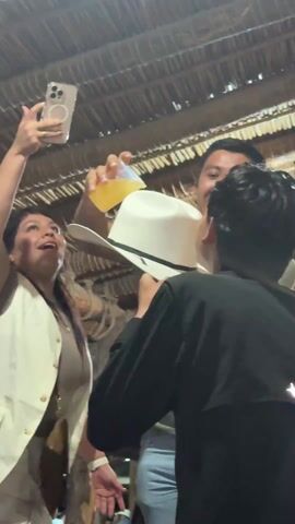 Here's a detailed description of the video:

The video shows a celebratory scene, likely at a party. A man wearing a white cowboy hat is pouring a yellow liquid (possibly beer or a mixed drink) from a glass into his hat. Another person in a black shirt is positioned to drink from the hat as it's being poured. To the left, a woman wearing a beige top and a white vest holds up a smartphone (likely an iPhone) to record the scene. She has her mouth open, indicating she's cheering or shouting in excitement.

The background suggests an outdoor or open-air venue, with a thatched or woven ceiling structure visible above. There's a bright light shining down on the scene, possibly from a spotlight or stage lighting, causing lens flare. Other people are partially visible in the background, suggesting a crowded gathering. The overall atmosphere seems lively and celebratory.