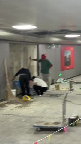 Three men are working on a construction site. One man in a red hard hat, green shirt, and light gray pants is holding a tool to a wall. Another man in a black shirt and black pants is squatting and working on something near the floor. A third man in a white t-shirt and black pants is also squatting and working on something near the floor. There are various tools and equipment scattered on the floor, including buckets, wires, and what appears to be a broom. In the background, there is a red sign with an image and text. The overall environment appears to be an indoor construction or renovation area.