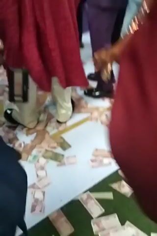 The video shows a gathering of people, many of whom are well-dressed. There are many bills of money strewn on the floor, and people are walking among them. A child is being given money directly by one of the men present. The overall impression is that this is a celebration with a generous display of wealth.