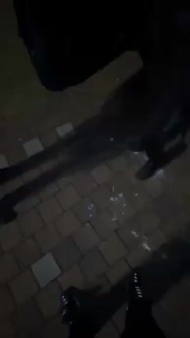 The video shows three individuals standing on a paved area outdoors at night. The ground is brick-like with some white liquid or substance scattered around. One person opens a car door and is wearing black shoes and pants. Another person is wearing a dark jacket and lighting something in their hand, possibly a cigarette. The third person is wearing a black hooded jacket and a cap, holding a cigarette, and is engaged in a conversation with the person lighting the cigarette. The environment appears to be a street or public area, with streetlights visible in the background.