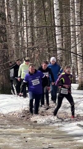 A group of runners is seen going through a watery and muddy patch of ground in a wooded area. Some snow is still present on the ground. The runners are wearing athletic clothing. They are wearing bibs with numbers on them. The trees are mostly bare.
