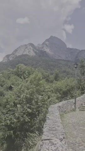 This is a landscape video set in Sargans, Switzerland. The scene features a majestic mountain dominating the background, with a lush green forest covering its lower slopes. In the foreground, a cobblestone path winds its way through the greenery, bordered by a rustic stone wall. A vintage-style lamppost stands tall beside the path, adding a touch of old-world charm.

The weather appears to be overcast, with a hazy sky and scattered clouds. The time of day is likely daytime, given the ambient light. The overall mood is serene and tranquil, inviting viewers to imagine a peaceful stroll through this scenic location. There is no visible text in the video.