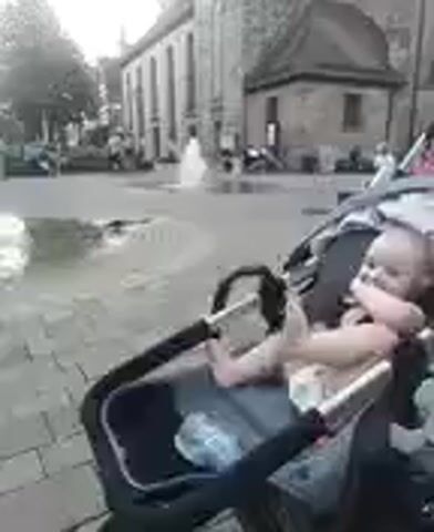 This video shows a baby sitting in a stroller in an outdoor square. The baby is wearing a diaper and a black tank top and is looking towards the camera with a slight smile. The stroller is dark gray and black with a black handle. A water bottle is visible inside the stroller.

In the background, there is a fountain with water spraying. Several people are walking around the square, and some children appear to be playing near the fountain. There are buildings in the background, including what looks like a church or old stone building. The ground is paved with cobblestones.

The overall mood of the video is relaxed and cheerful, with the baby appearing content. The lighting suggests it is daytime, possibly late afternoon due to the warm tones in the sky. The setting appears to be a public square in a European town, likely Altdorf bei Nürnberg, Germany, as indicated by the context. There is no visible text in the video.