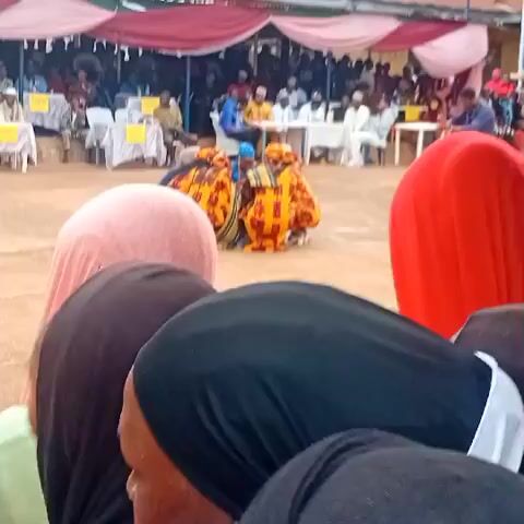 A ground-level video shows a group of people, with their backs to the camera, watching an outdoor event. In the foreground, the backs of people's heads are visible, covered by hijabs in shades of pink, black, and red. Beyond them, in a sandy clearing, a group of four individuals in bright yellow and orange patterned outfits are seated on the ground, seemingly performing or participating in an activity. Further back, under a large tent with red and white draping, many people are seated at tables and chairs, observing the event. Another person in a light yellow traditional outfit is standing near the performing group, while others stand further in the background. The ground is a light brown, sandy color, and the overall atmosphere suggests a communal gathering or celebration.